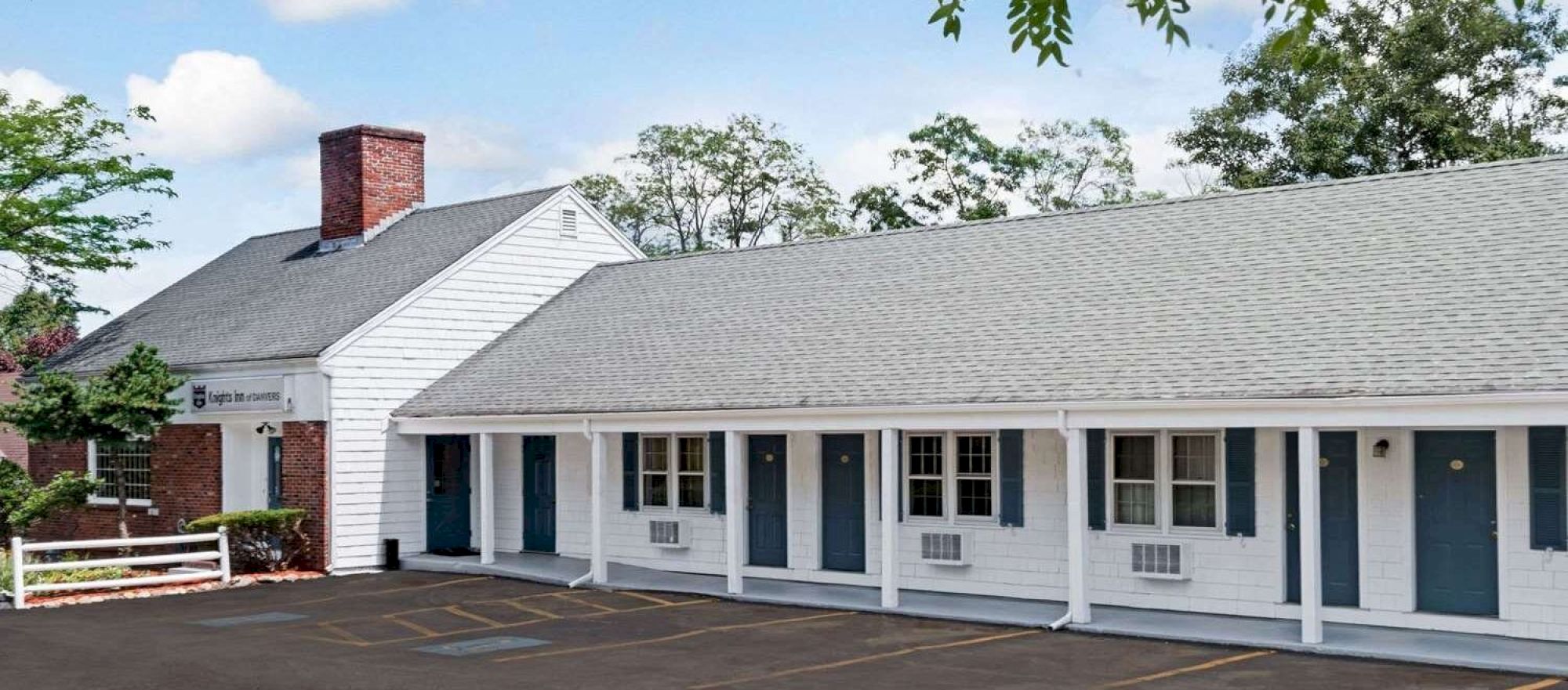 A row of single-story white buildings with dark doors and shutters, a parking lot in front, and a clear blue sky with green trees overhead, ending now.