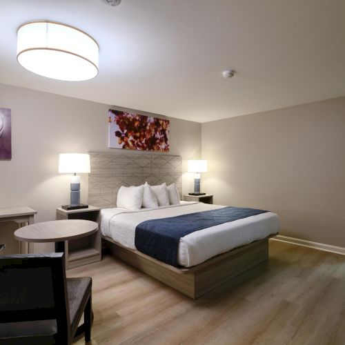 A modern hotel room with a king bed, two nightstands with lamps, a seating area, and wall art; doorway to a small closet area on the right.