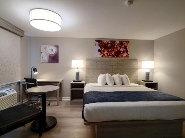 A modern hotel room with a king bed, two nightstands, lamps, artwork, a small table and chairs, and a desk area by the window.