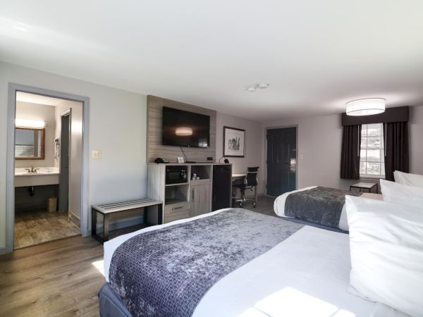 A hotel room with two large beds, a TV above a minibar/console, a small desk, and an open doorway to a separate bathroom area, bright and spacious.