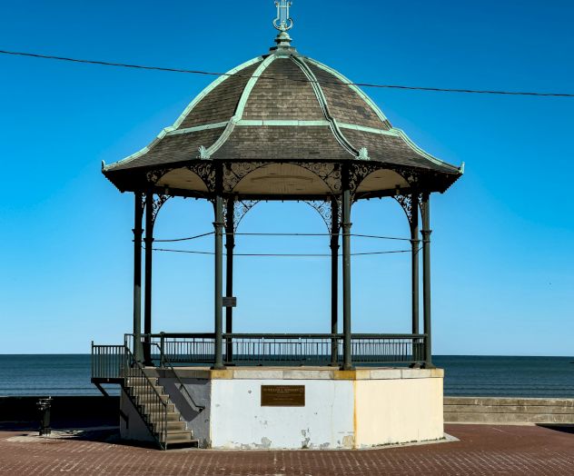 A seaside gazebo with a domed, ornate metal roof and open sides on a brick promenade by the water, inviting views over the sea.
