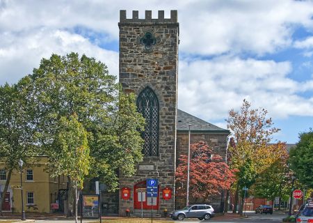 A tall stone church with a tower, Gothic window, surrounded by trees and a small town street with parked cars.